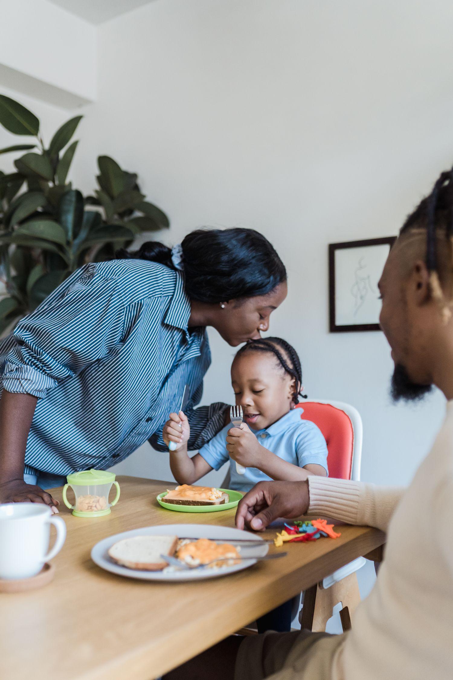A mother kissing her young son on the head at the family kitchen table while he eats breakfast