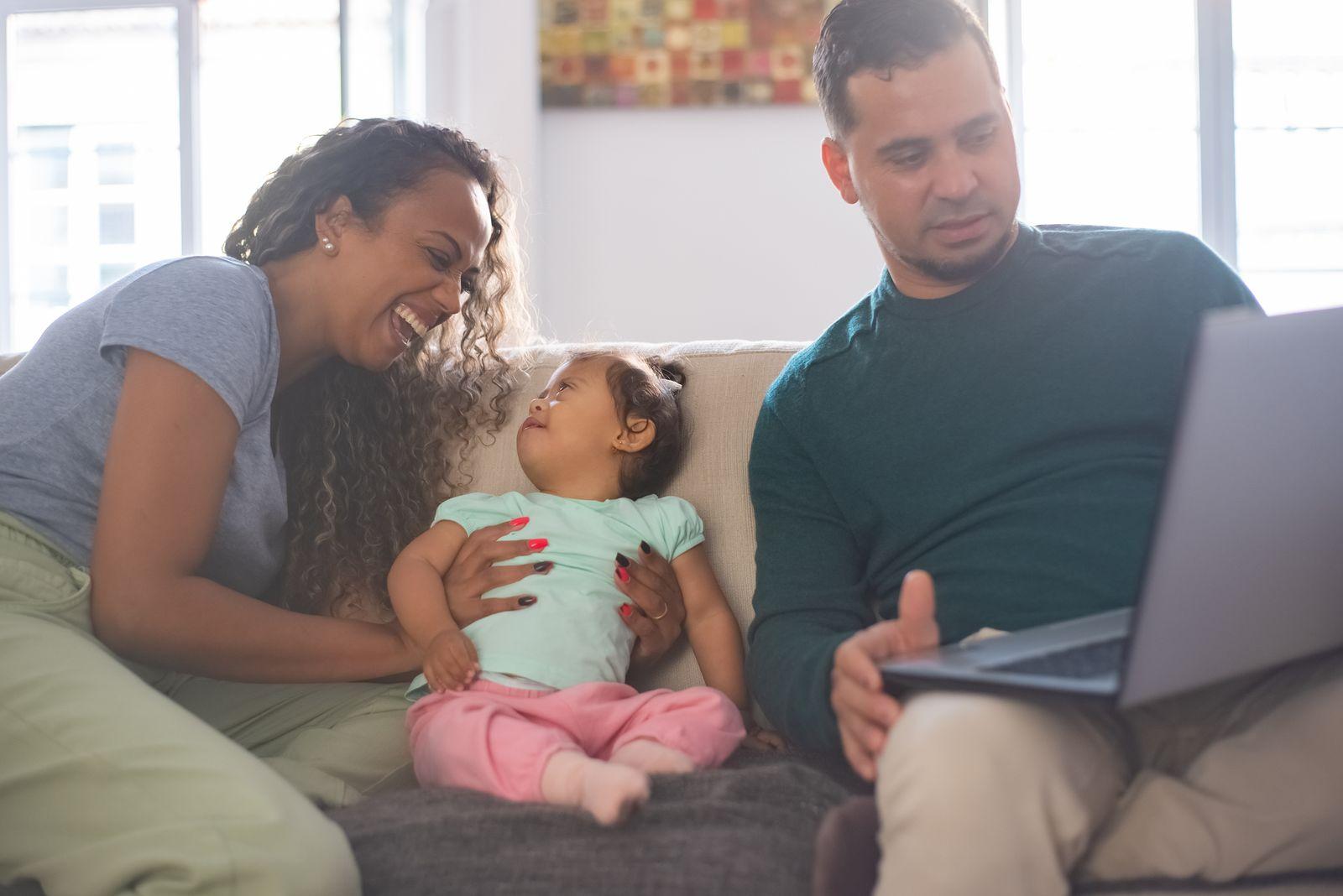 A mother laughing with her young child on the couch at home while a parent works nearby